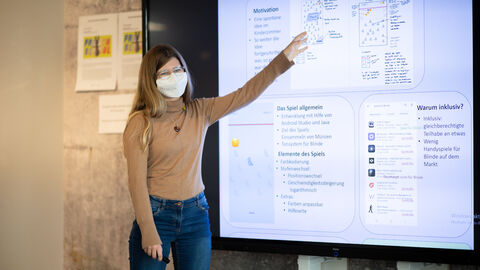 Lilly Schwarz präsentiert das Konzept ihrer App. Sie zeigt auf einen großen Bildschirm und lächelt unter ihrer Maske