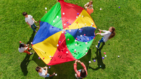 Aufsicht auf Kinder, die um ein rundes, buntes Tuch verteilt stehen, auf dem Bälle herumfliegen.