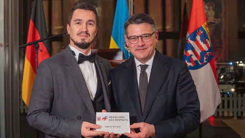 Ministerpräsident Boris Rhein und Evgenij Voznyuk stehen vor Deutschlandflagge, Ukraine-Flagge und Hessen-Flagge. Beide halten Auszeichnung mit dem Logo "Menschen des Respekts"