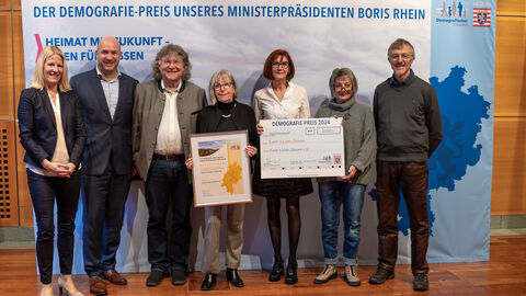 Eine Gruppe Personen steht vor einem Aufsteller mit der Aufschrift "Der Demografie-Preis unseres Ministerpräsidenten Boris Rhein". Eine Person hält eine gerahmte Urkunde in Händen, eine weitere einen Scheck.